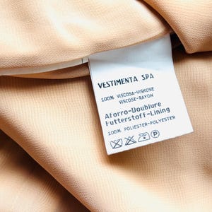 May include: Close-up of a beige fabric with a tag. The tag reads "VESTIMENTA SPA" and lists fabric composition: 100% Viscose-Rayon for the main fabric and 100% Polyester for the lining. The fabric has a subtle texture.
