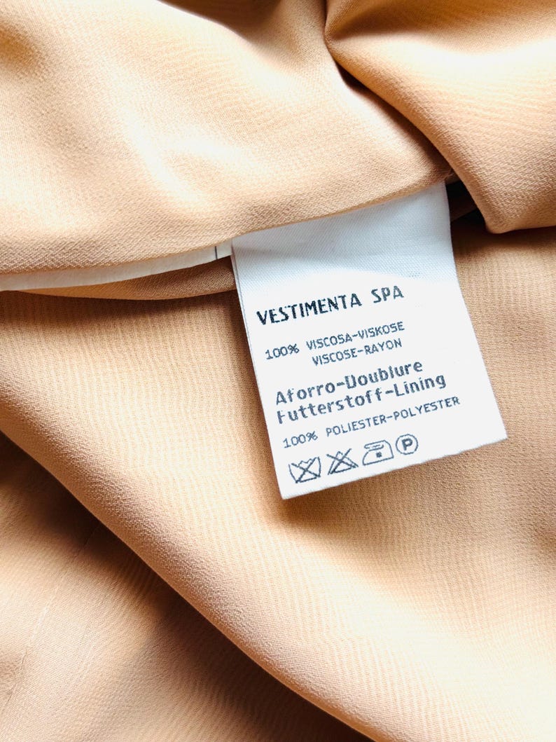 May include: Close-up of a beige fabric with a tag. The tag reads "VESTIMENTA SPA" and lists fabric composition: 100% Viscose-Rayon for the main fabric and 100% Polyester for the lining. The fabric has a subtle texture.