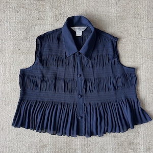 May include: A sleeveless navy blue blouse with a collared neckline and button-down front. The top features vertical textured detailing across the chest and a pleated peplum hem. The label reads "VIVIENNE TAM".