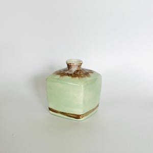 May include: A small, square ceramic vase with a light green glaze and a brown band around the top. The vase has a slightly rounded top.