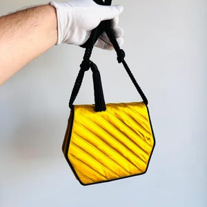 May include: A yellow satin handbag with a geometric shape and black trim. The bag features diagonal stitching and a black rope handle. The bag is held by a gloved hand.