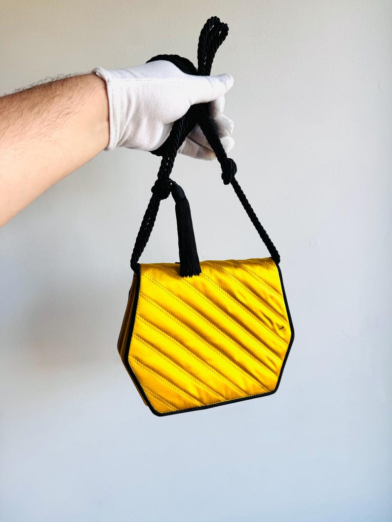 May include: A yellow satin handbag with a geometric shape and black trim. The bag features diagonal stitching and a black rope handle. The bag is held by a gloved hand.