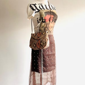 K&ouml;nnte beinhalten: Eine Schaufensterpuppe zeigt ein cremefarbenes Grafik-T-Shirt, einen braunen Rock mit silbernen Verzierungen und eine kleine Crossbody-Tasche aus Schlangenleder. Das T-Shirt hat eine schwarz-rote Grafik. Der Rock hat einen transparenten &Uuml;berzug.
