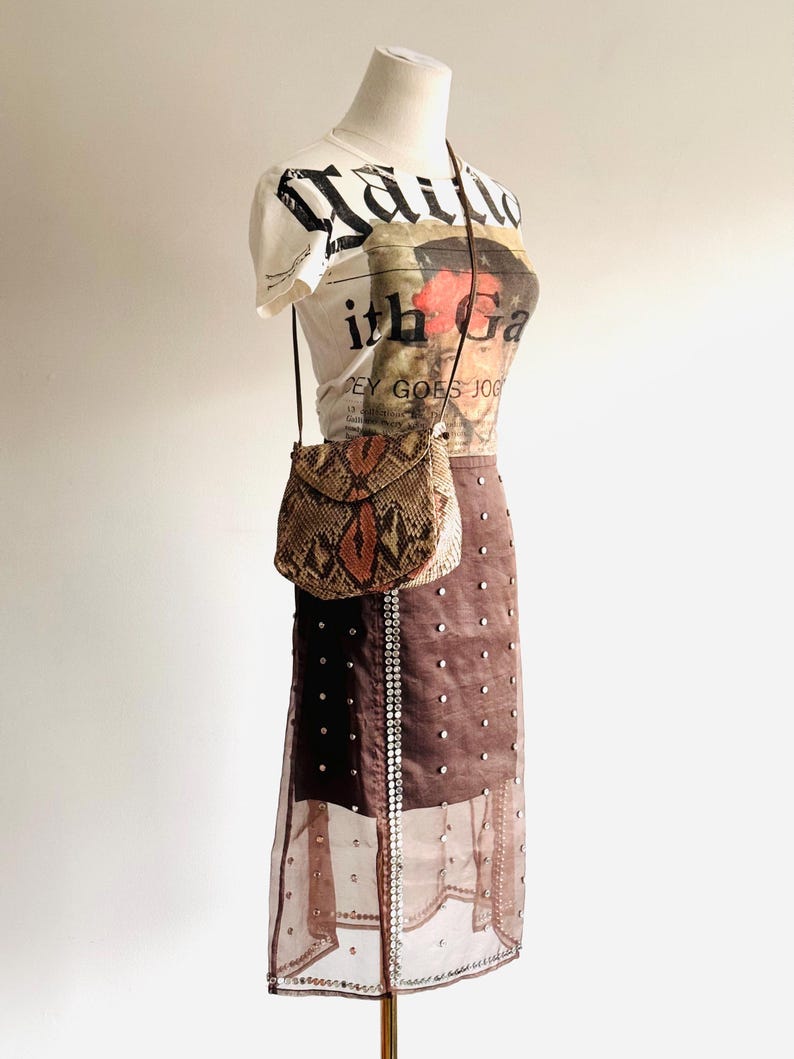 K&ouml;nnte beinhalten: Eine Schaufensterpuppe zeigt ein cremefarbenes Grafik-T-Shirt, einen braunen Rock mit silbernen Verzierungen und eine kleine Crossbody-Tasche aus Schlangenleder. Das T-Shirt hat eine schwarz-rote Grafik. Der Rock hat einen transparenten &Uuml;berzug.