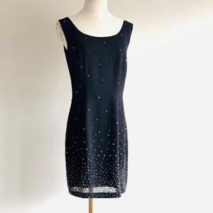 May include: A sleeveless black knee-length dress featuring a scoop neckline and a fitted silhouette. The fabric is adorned with scattered silver-toned beads that become increasingly dense toward the hem, creating a gradient effect throughout.
