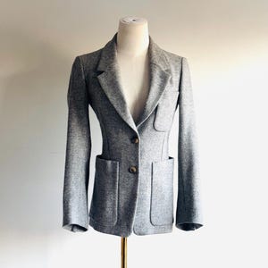 May include: A gray blazer with two front pockets and two brown buttons. The blazer is displayed on a mannequin. The blazer has a tailored fit and a classic lapel design. The fabric appears to be a wool blend.
