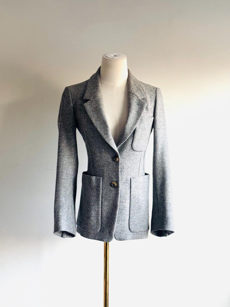 May include: A gray blazer with two front pockets and two brown buttons. The blazer is displayed on a mannequin. The blazer has a tailored fit and a classic lapel design. The fabric appears to be a wool blend.