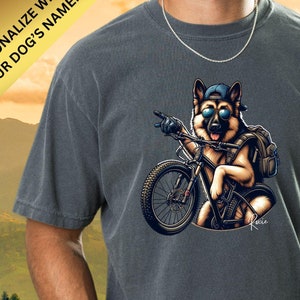 May include: A dark gray t-shirt with a cartoon image of a German Shepherd dog wearing sunglasses and a blue baseball cap, riding a mountain bike. The dog is wearing a backpack and the text "Pepper" is on the bottom right corner of the shirt.