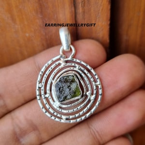 May include: A silver pendant with a green gemstone set in a circular design. The pendant has a textured, concentric circle pattern around the gemstone.