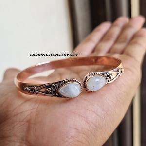 Puede incluir: Brazalete abierto de color cobre con dos piedras claras en forma de pera. El brazalete tiene filigranas decorativas y el texto "EARRINGJEWELLRYGIFT".