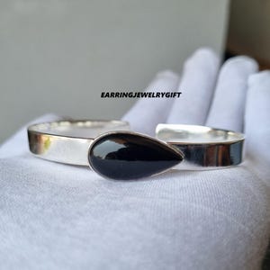 May include: A silver cuff bracelet with a teardrop-shaped black stone. The bracelet is displayed on a white surface. The text "EARRINGJEWELRYGIFT" is visible.