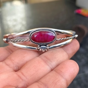 Könnte beinhalten: Silbernes Armreif-Armband mit einem großen, ovalen Rubin-Edelstein. Der Rubin ist in einer detaillierten Lünette gefasst, umgeben von einem gedrehten Seildesign und dekorativen Akzenten. Das Armband wird in einer Hand gehalten.