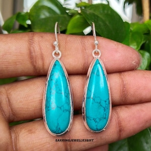 May include: A pair of turquoise teardrop dangle earrings with silver settings. The earrings are held in a hand against a green leafy background.
