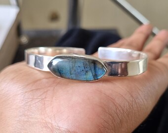 May include: A silver cuff bracelet with a large, oval labradorite stone displaying blue and green iridescence. The bracelet has a simple, polished design, highlighting the gemstone. A piece of jewellery.