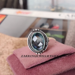 Pode incluir: Um anel de prata com uma grande pedra preciosa oval facetada de cor azul claro. O anel tem um design decorativo de corda torcida em torno da pedra preciosa.