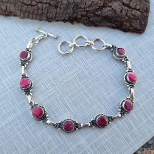 以下が含まれることがあります： トグル留め具と7つの丸いピンクの宝石が付いたシルバーブレスレット。ブレスレットはスターリングシルバー製で、繊細なチェーンのデザインが特徴です。