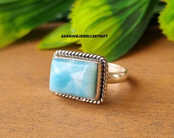 May include: A silver ring with a rectangular, light blue gemstone. The gemstone is set in a decorative, rope-like border. The ring is on a wooden surface. The text "earringjewellerygift" is visible.