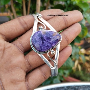 Pode incluir: Um bracelete de prata com uma pedra preciosa roxa e dourada. O bracelete tem um design delicado com volutas e uma pedra em forma de lágrima.