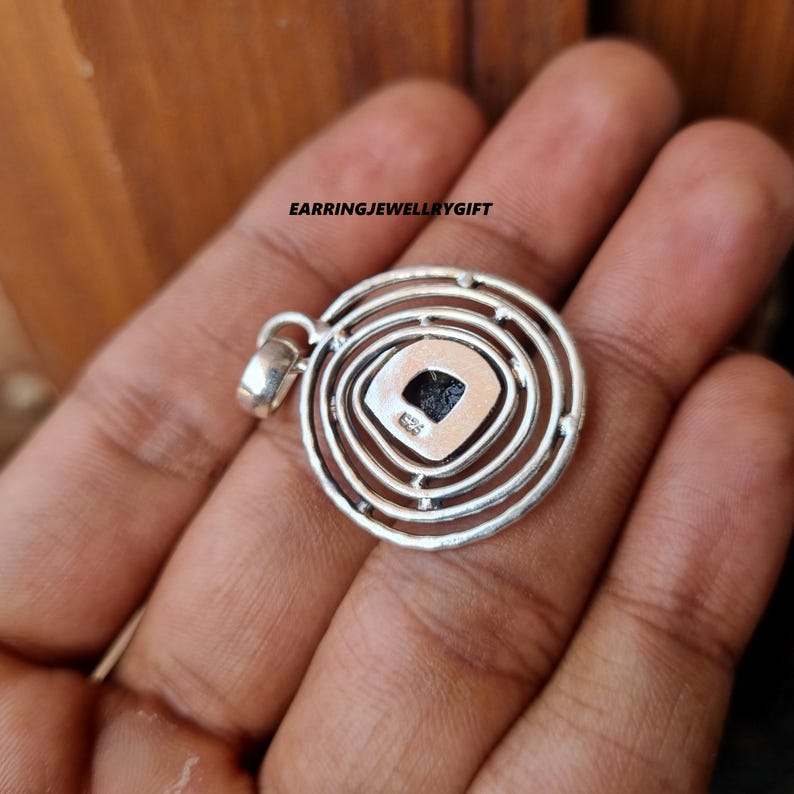 May include: Silver pendant with a circular design featuring concentric circles and a square-shaped gemstone in the center. The pendant has a bail for attaching to a chain.