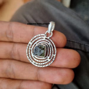 May include: A silver pendant with a green gemstone set in a circular design with concentric circles. The pendant is on a hand.