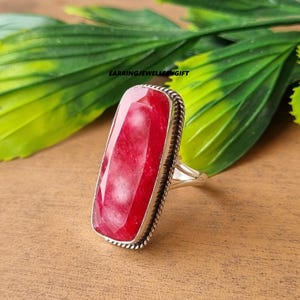 May include: A silver ring with a large, rectangular, faceted red gemstone. The gemstone is set in a silver band with a textured border. The ring is displayed on a wooden surface with green leaves in the background. The text "EARRINGJEWELLERYGIFT" is visible.