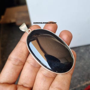 Puede incluir: Un colgante ovalado negro con una anilla de plata. El colgante está hecho de una piedra lisa y pulida.
