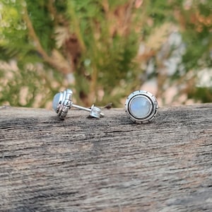 May include: A pair of silver earrings with a round moonstone gemstone in each. The earrings have a textured silver border around the stone.