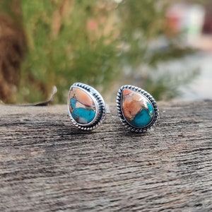 May include: A pair of teardrop-shaped stud earrings. Each earring features a vibrant combination of orange, turquoise, and silver. The stones are set in a silver frame with a twisted rope design around the edge. The earrings are displayed on a wooden surface.