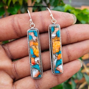 May include: A pair of silver dangle earrings with a rectangular turquoise, orange, and white stone inlay. The earrings have a rope-like border around the stone.