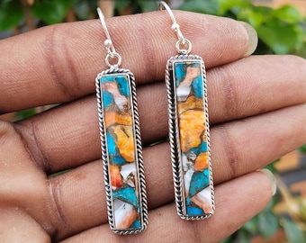 May include: A pair of silver dangle earrings with a rectangular turquoise, orange, and white stone inlay. The earrings have a rope-like border around the stone.