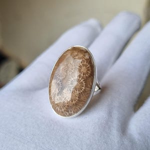 Puede incluir: Anillo ovalado con una piedra de coral fosilizado y una banda de plata. El coral presenta un patrón natural detallado en tonos marrones y crema. El diseño del anillo es moderno y elegante. Una joya única.