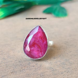 May include: A silver ring featuring a large, teardrop-shaped, faceted ruby gemstone. The ruby is a vibrant shade of pink-red. The ring is set on a wooden surface. The text "EARRINGJEWELLERYGIFT" is visible in the image.