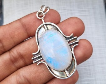 May include: A silver pendant featuring an oval moonstone with a blue and white iridescent glow. The stone is set in a decorative silver frame with an ornate bail for a necklace chain. The pendant is held in a hand.