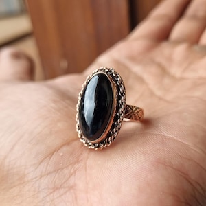 May include: A copper-colored ring with an oval, glossy black stone. The stone is set in a decorative frame with a braided rope design. The ring's band features an etched floral pattern. The ring is displayed on a hand.