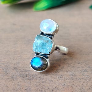 Puede incluir: Un anillo de plata con tres piedras preciosas: una piedra lunar redonda de color azul pálido, una aguamarina azul claro en bruto y un labradorita iridiscente redondo. El anillo está sobre una superficie de madera.