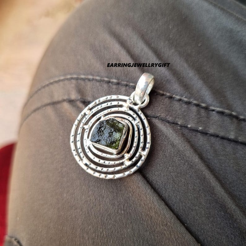 May include: A silver pendant with a green gemstone set in a circular design. The pendant has a delicate, intricate pattern and is perfect for adding a touch of elegance to any outfit.