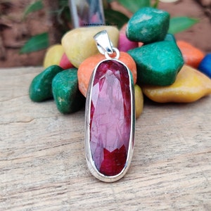 Può includere: Un ciondolo con una pietra preziosa rossa incastonata in argento. La pietra preziosa è di forma ovale e ha una sfaccettatura. Il ciondolo è appeso a una catena d'argento.