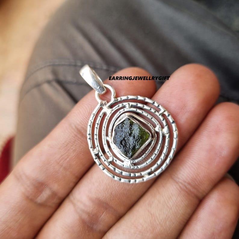 May include: A silver pendant with a green gemstone set in a circular design with concentric circles. The pendant is on a hand.