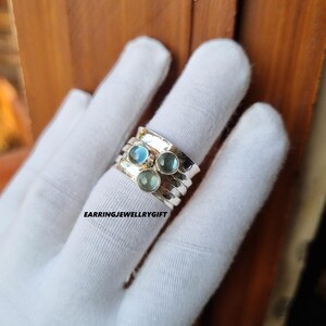 Può includere: Un anello d'argento con tre pietre rotonde, azzurre e verdi. L'anello ha una fascia strutturata e viene indossato su un dito. Il testo "EARRINGJEWELLRYGIFT" è visibile.