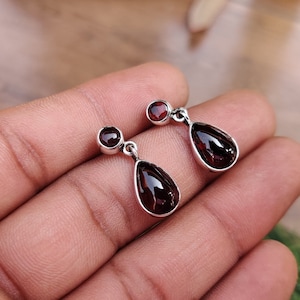 Può includere: Un paio di orecchini pendenti in argento con gocce di pietre preziose rosse. Gli orecchini presentano un piccolo gioiello rotondo nella parte superiore e un gioiello più grande a forma di lacrima sotto.