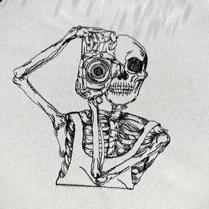 May include: Black and white embroidery art on white fabric, featuring a skeleton with a camera lens for a head. The embroidery is set in a round hoop, with a dark grey background.