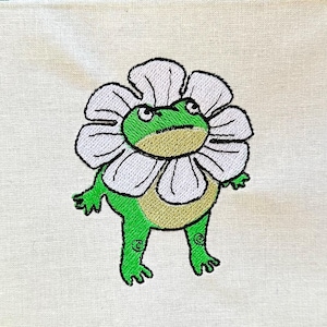 Peut inclure: Patch brodé d'une grenouille verte portant une fleur blanche sur un fond de tissu blanc.