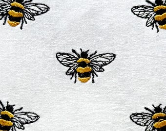 May include: Embroidered hoop art featuring six black and yellow bees on a white background. The hoop has a gold chain border with gold dots around the edge.