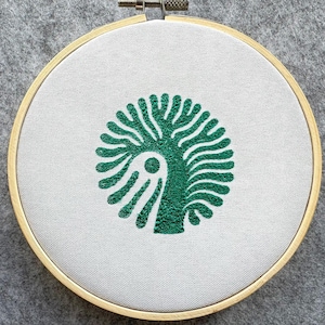 Könnte beinhalten: Ein grünes gesticktes Baumdesign auf einem weißen Stoff, der über einen hölzernen Stickrahmen gespannt ist.