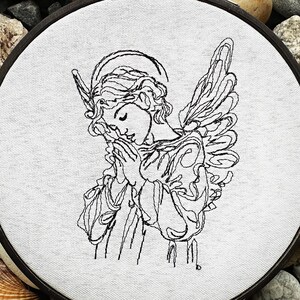 Pode incluir: Bordado de linha preto e branco de um anjo com asas, rezando com as mãos juntas, sobre um fundo de tecido branco.