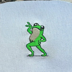 May include: Embroidered green frog with a white background. The frog is dancing with its arms raised and one leg bent.