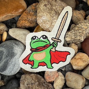 Könnte beinhalten: Gestickter Aufnäher mit einem grünen Frosch, der einen roten Umhang trägt und ein Schwert hält. Der Frosch hat große Augen und befindet sich vor einem Hintergrund aus Steinen und Kieselsteinen. Der Aufnäher hat ein skurriles, cartoonartiges Design.