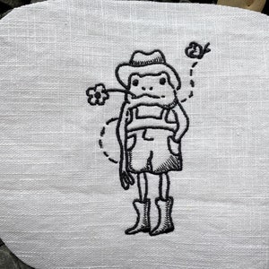 May include: Black and white embroidered patch of a frog wearing a hat and overalls, holding a flower and with a bird flying above.