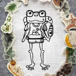May include: A black and white embroidered design of a frog wearing glasses and a backpack on a white fabric background. The frog is surrounded by a circle of various stickers, including a bear, a rabbit, and plants.