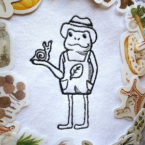 Peut inclure: Un dessin au trait noir et blanc d'une grenouille portant un chapeau et une salopette tenant un escargot. La grenouille est entourée de divers autocollants sur un fond blanc.